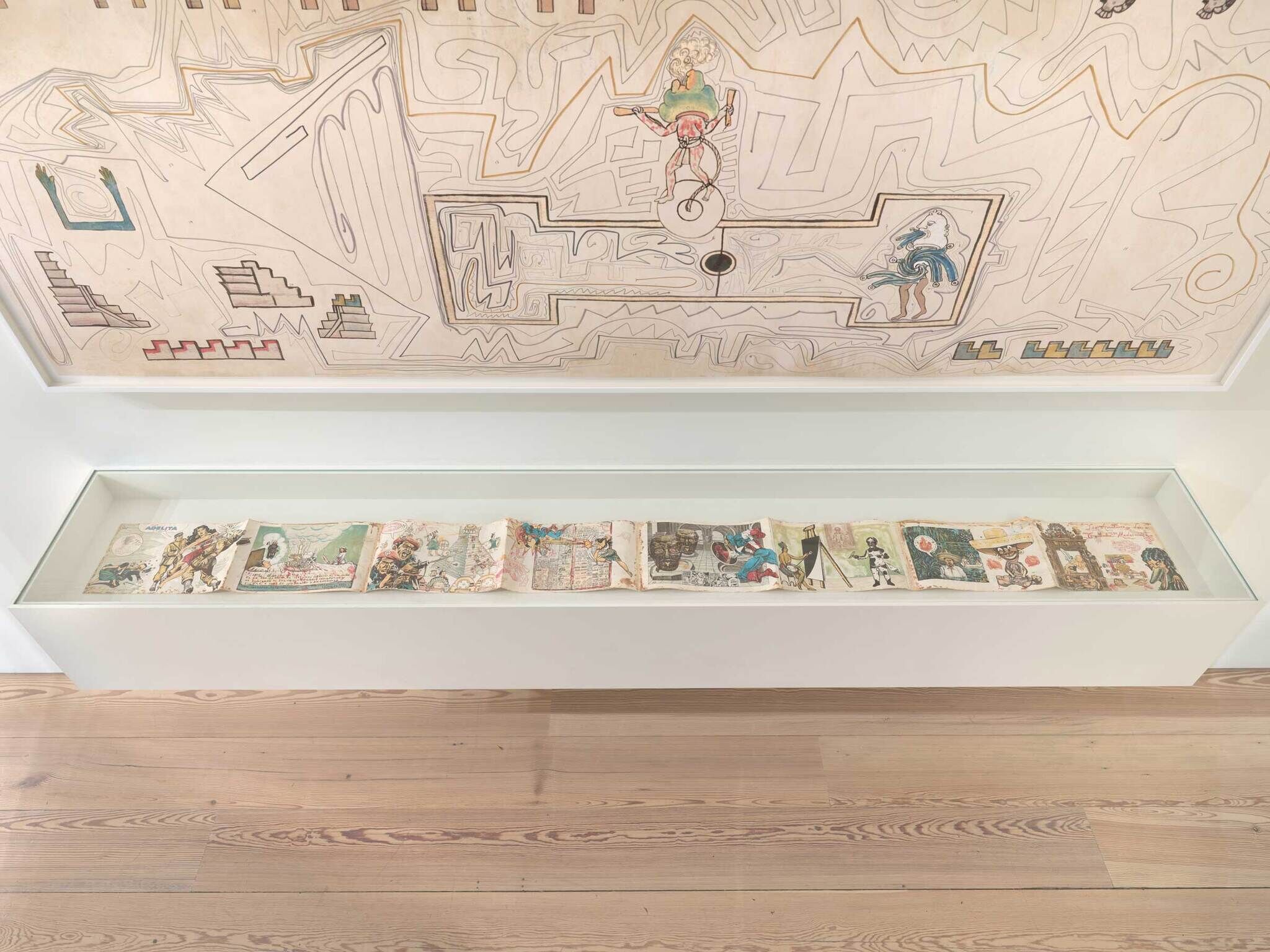 A long illustrated accordion book displayed under a large abstract mural drawing in a gallery.