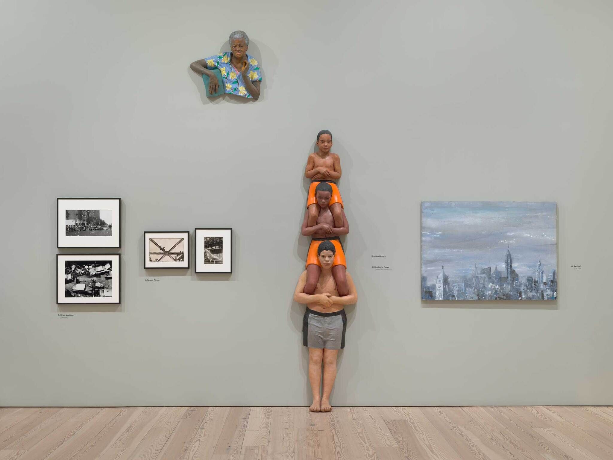 Four life-size figures stacked vertically against a gallery wall, with a painted head above them.