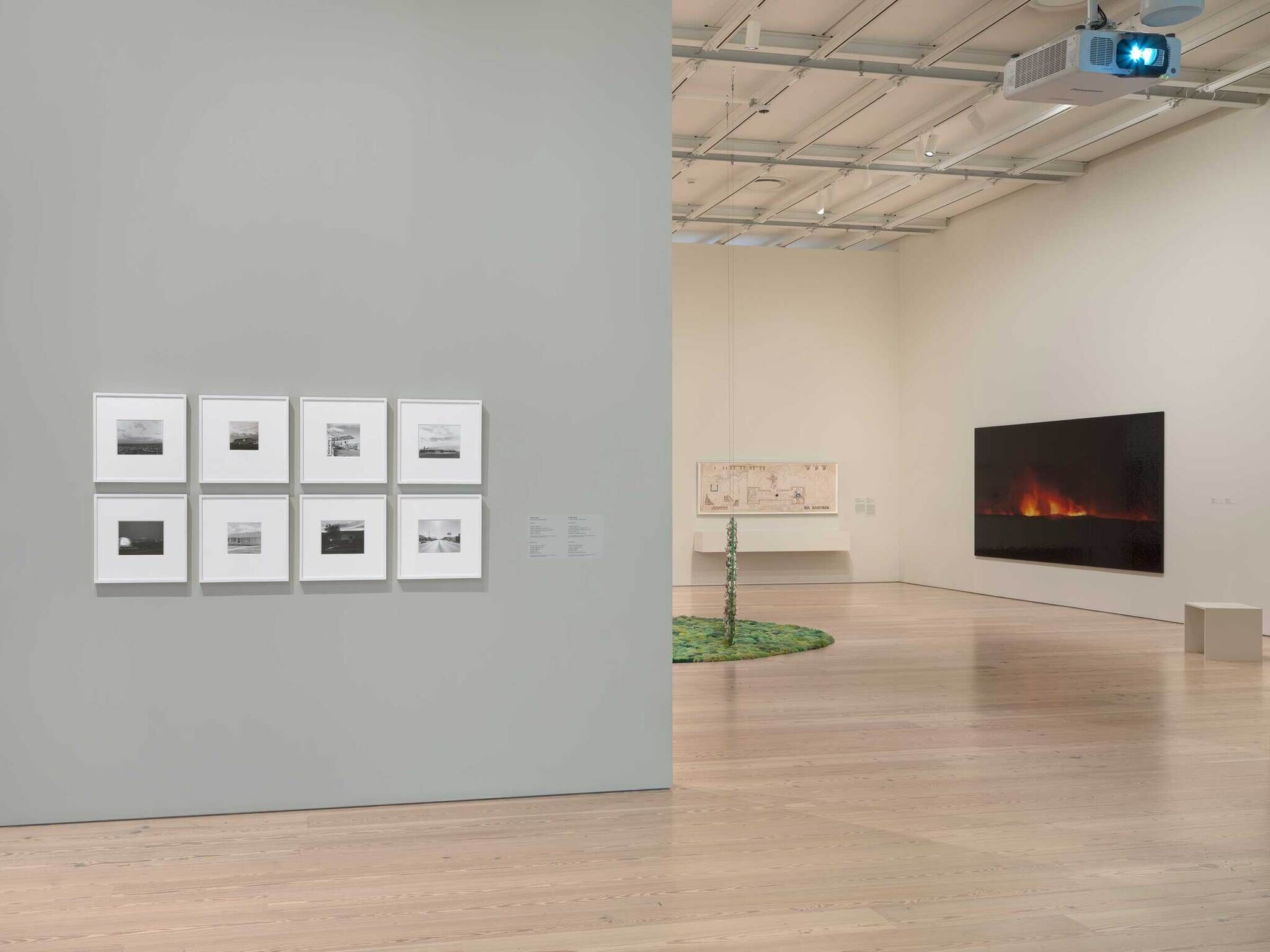 Nine framed black-and-white photographs hang on a wall beside a gallery room with a fiery video.