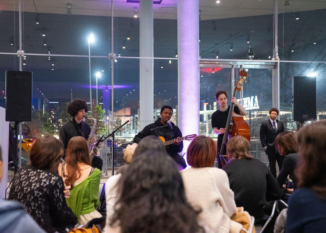 A small jazz band performs with saxophone, guitar, and double bass for a seated audience.
