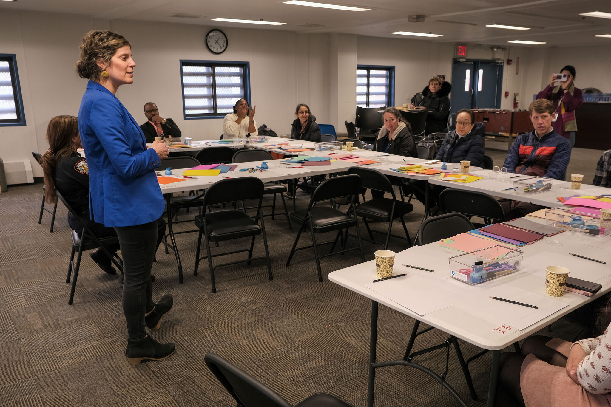 Woman in blue blazer leads a workshop while participants sit around tables with art supplies.