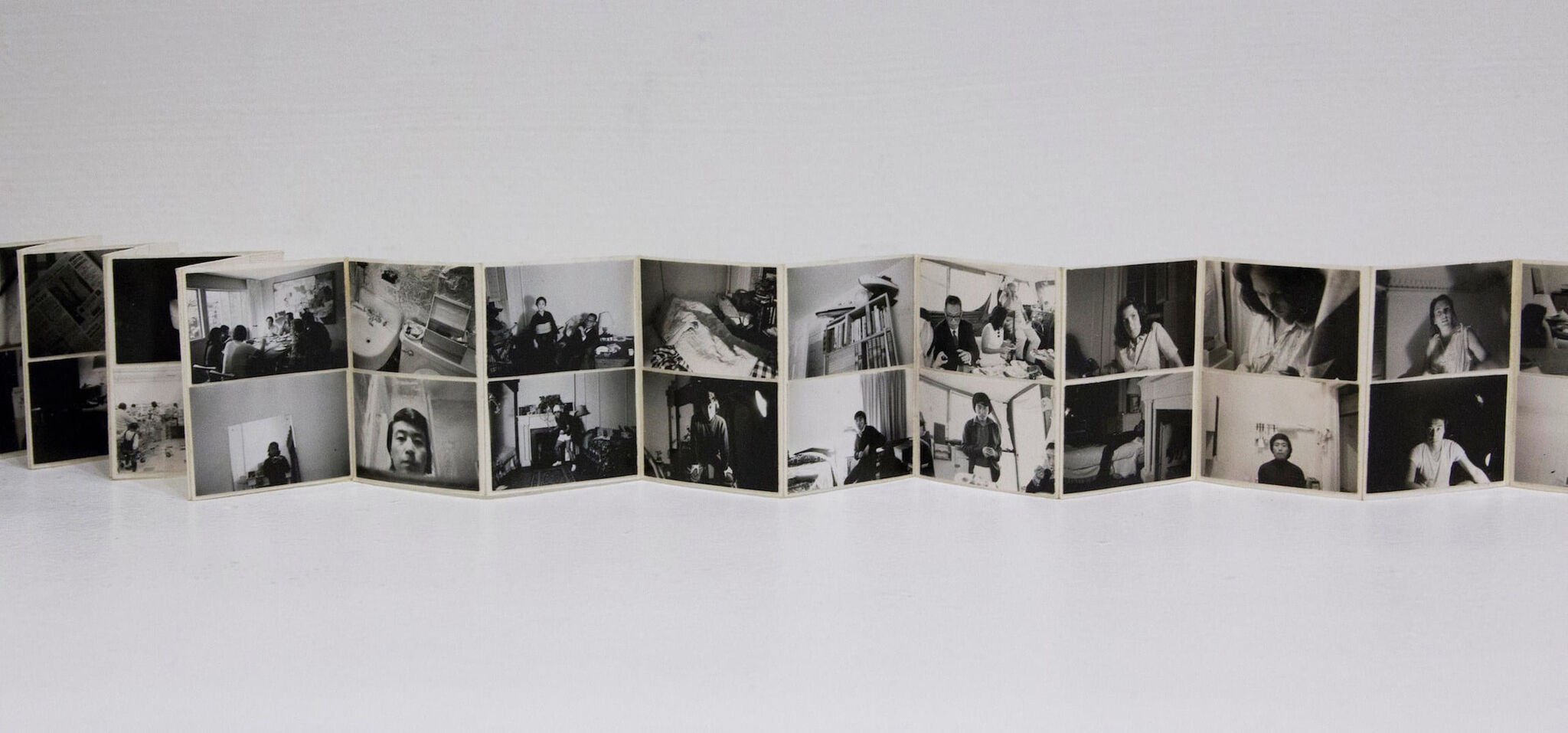 A long accordion-style photo book displayed fully extended on a white surface. Each panel features small black-and-white portraits and interior scenes.