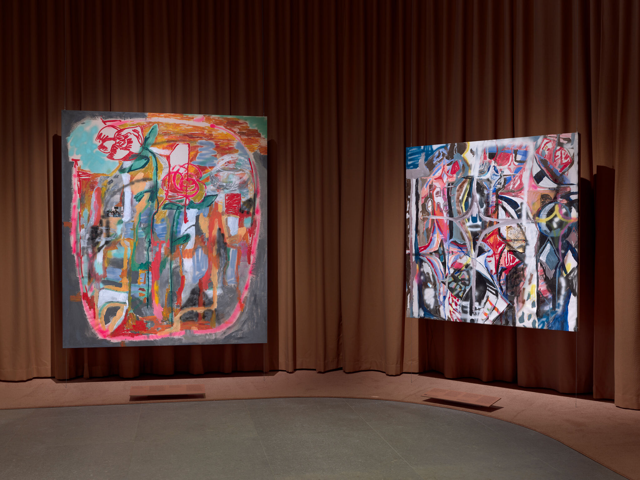 Two large colorful abstract paintings hang side by side against a brown curtain backdrop in a gallery.