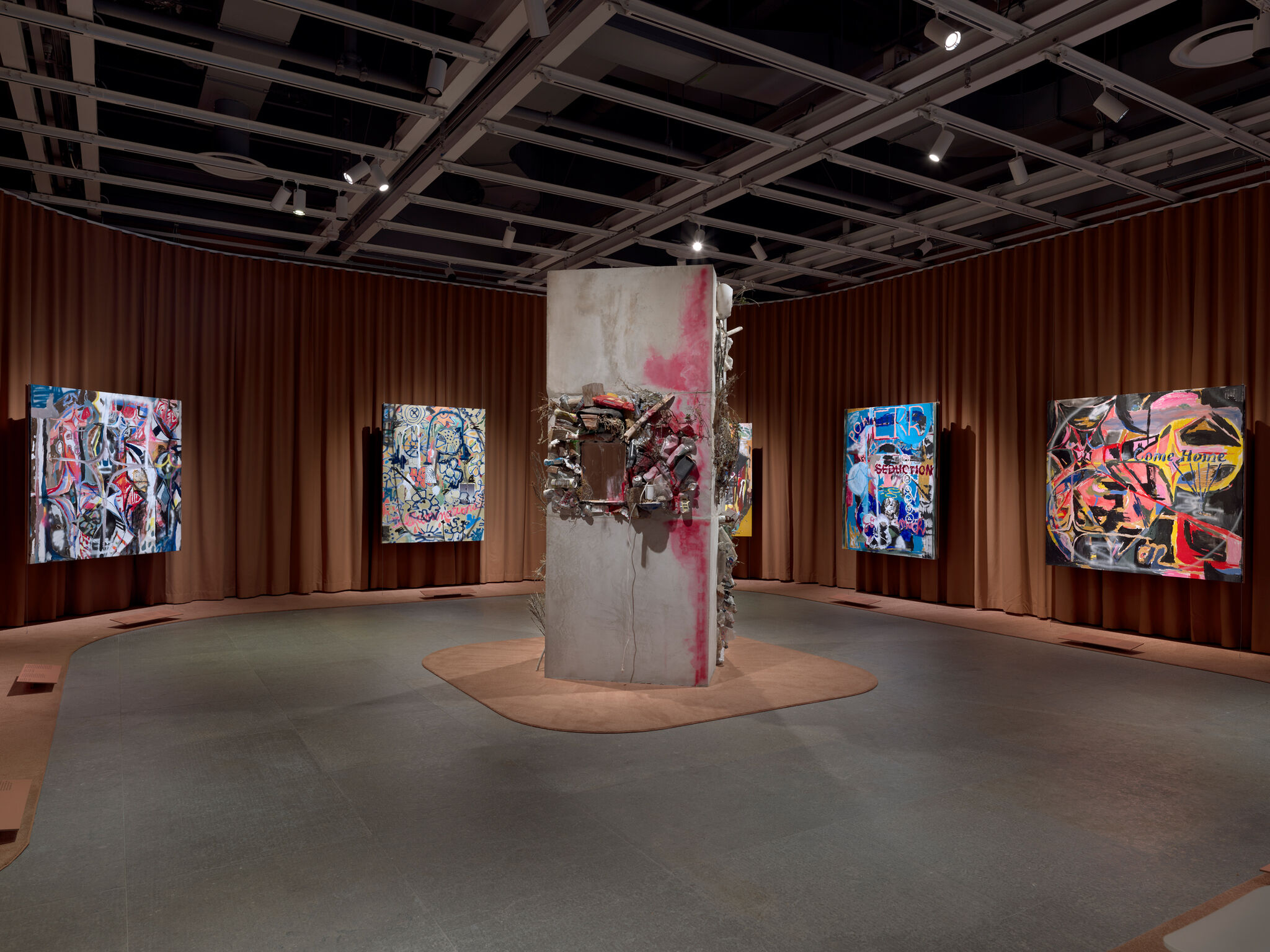 Sculptural pillar with attached objects stands in the center of a gallery surrounded by abstract, colorful paintings.