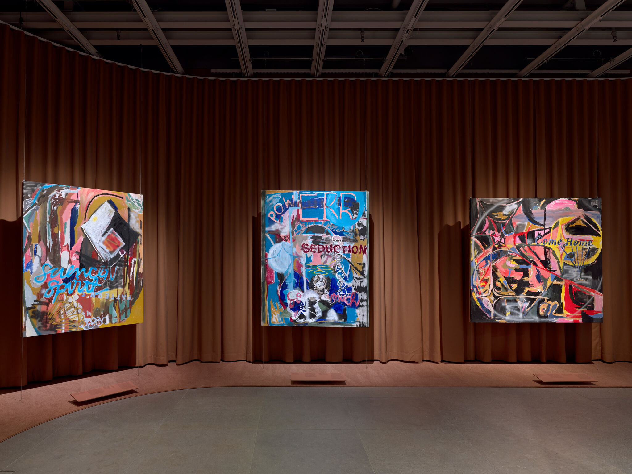 Three large colorful abstract paintings hang on a brown curtained wall in a gallery.
