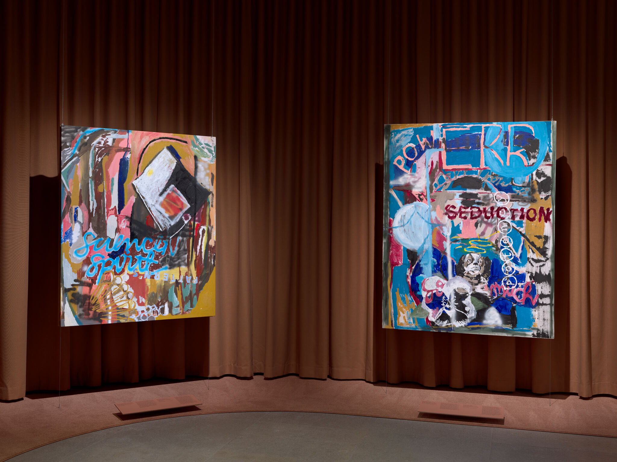 Two large colorful abstract paintings hang from wires in a gallery space with brown curtains.