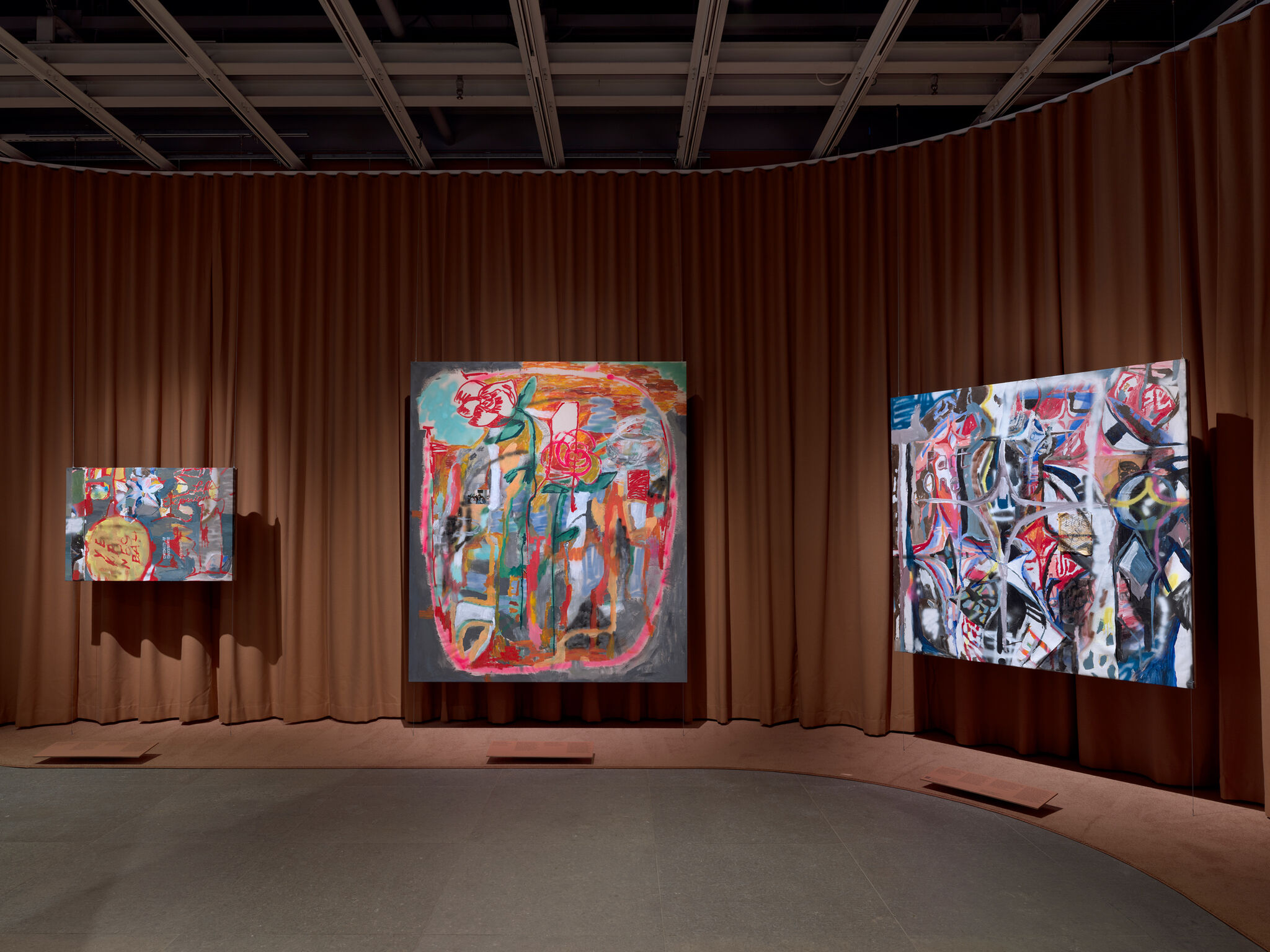 Three large colorful abstract paintings hang on a brown curtain wall in a gallery.