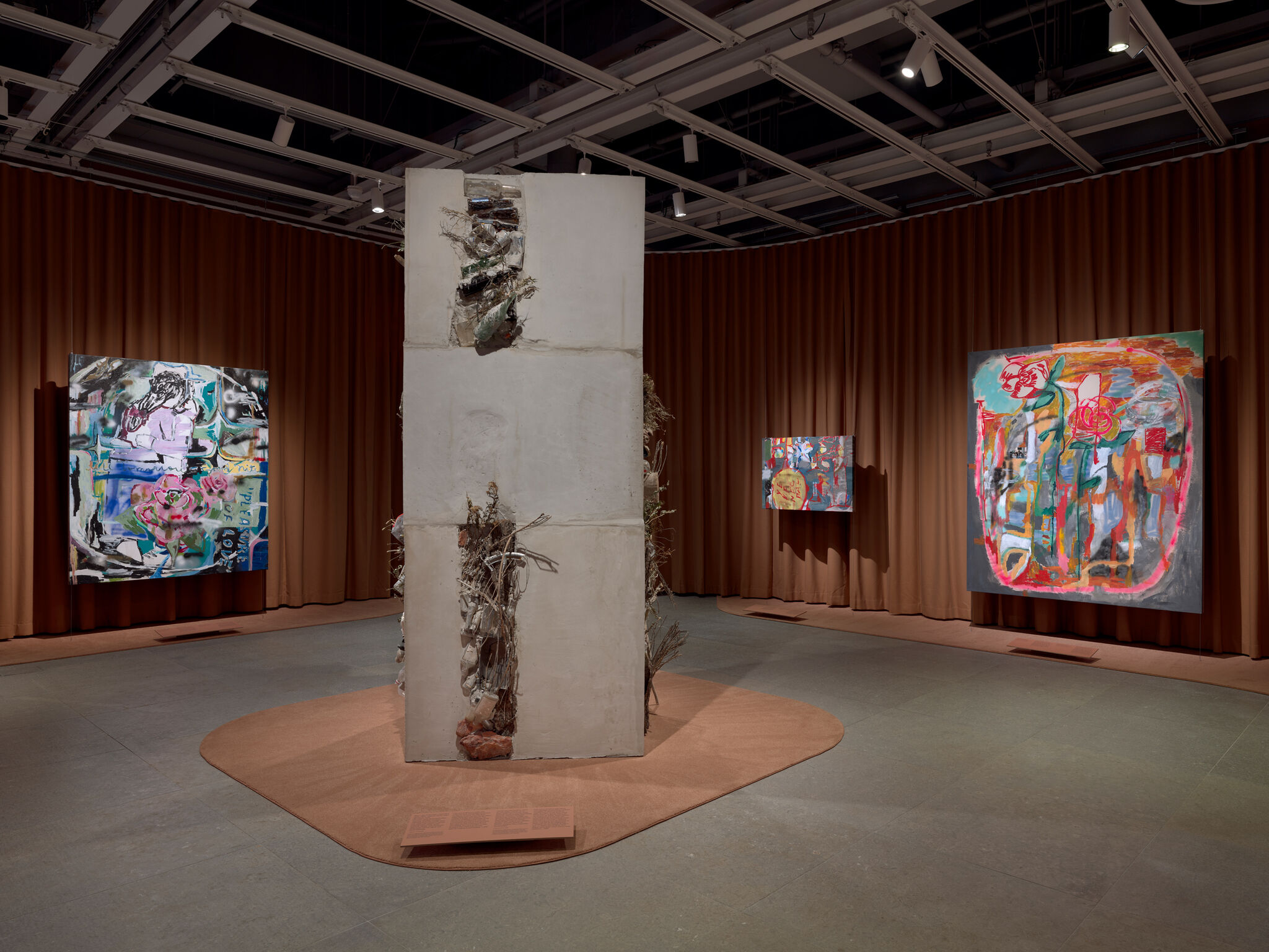 A tall concrete sculptural pillar with embedded debris stands amid colorful abstract paintings in a gallery.