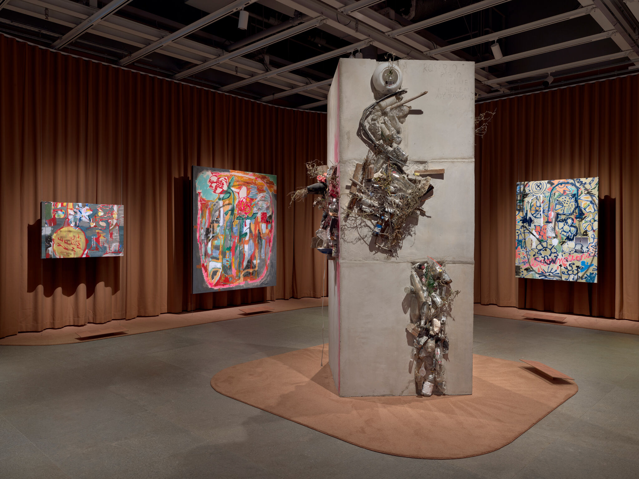 Concrete pillar sculpture covered in found objects stands in a gallery surrounded by colorful abstract paintings.