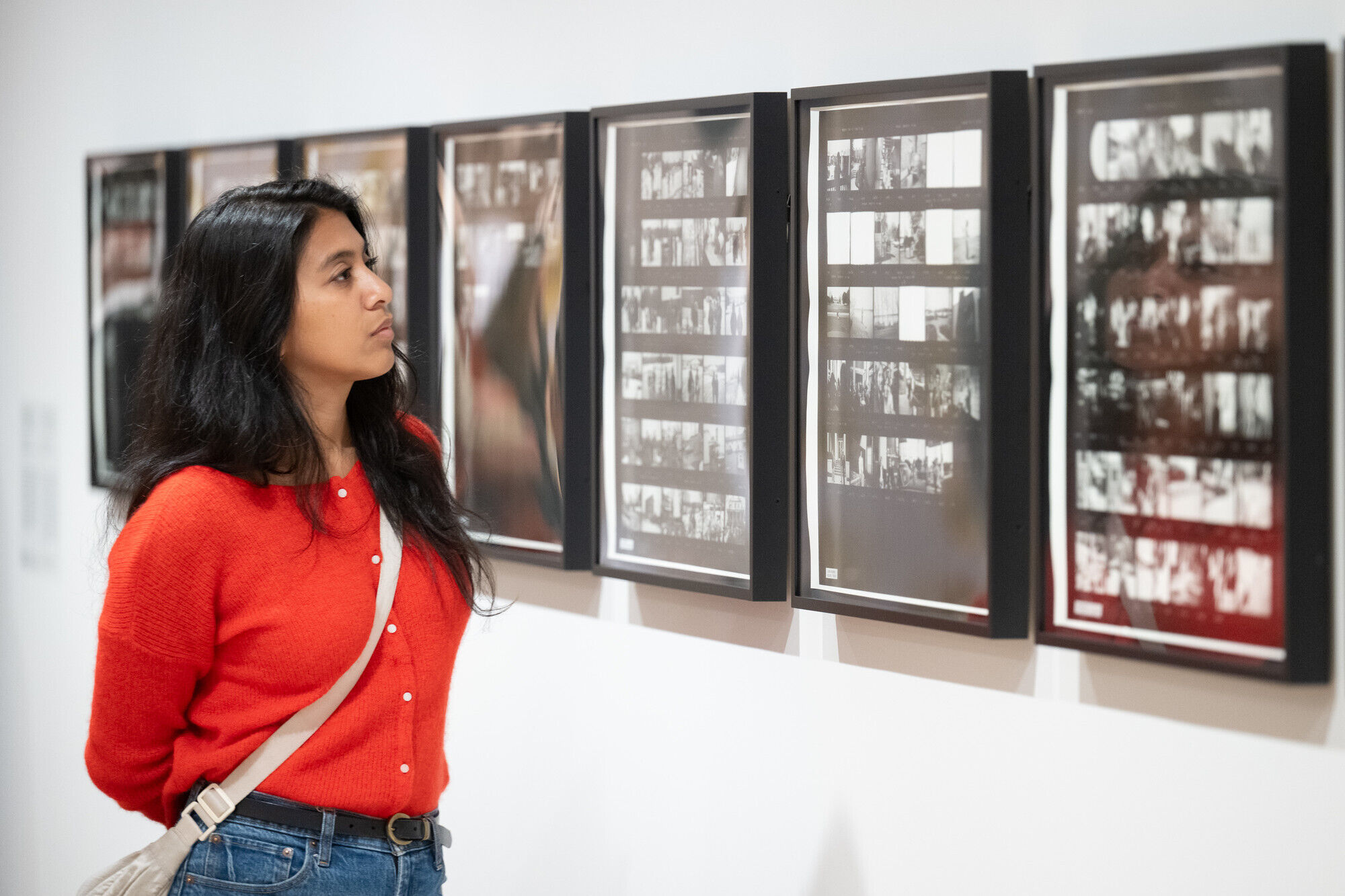 A woman in a red sweater studies framed photographic contact sheets displayed on a gallery wall.