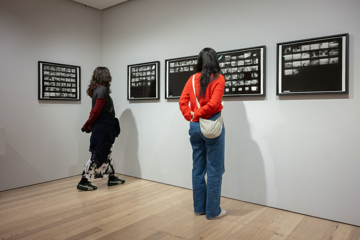 Two people stand in a gallery closely examining framed black-and-white contact sheet photographs on the wall.