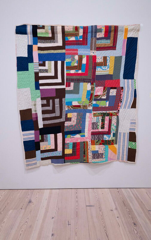 A colorful patchwork quilt with geometric log-cabin blocks hangs on a white gallery wall.