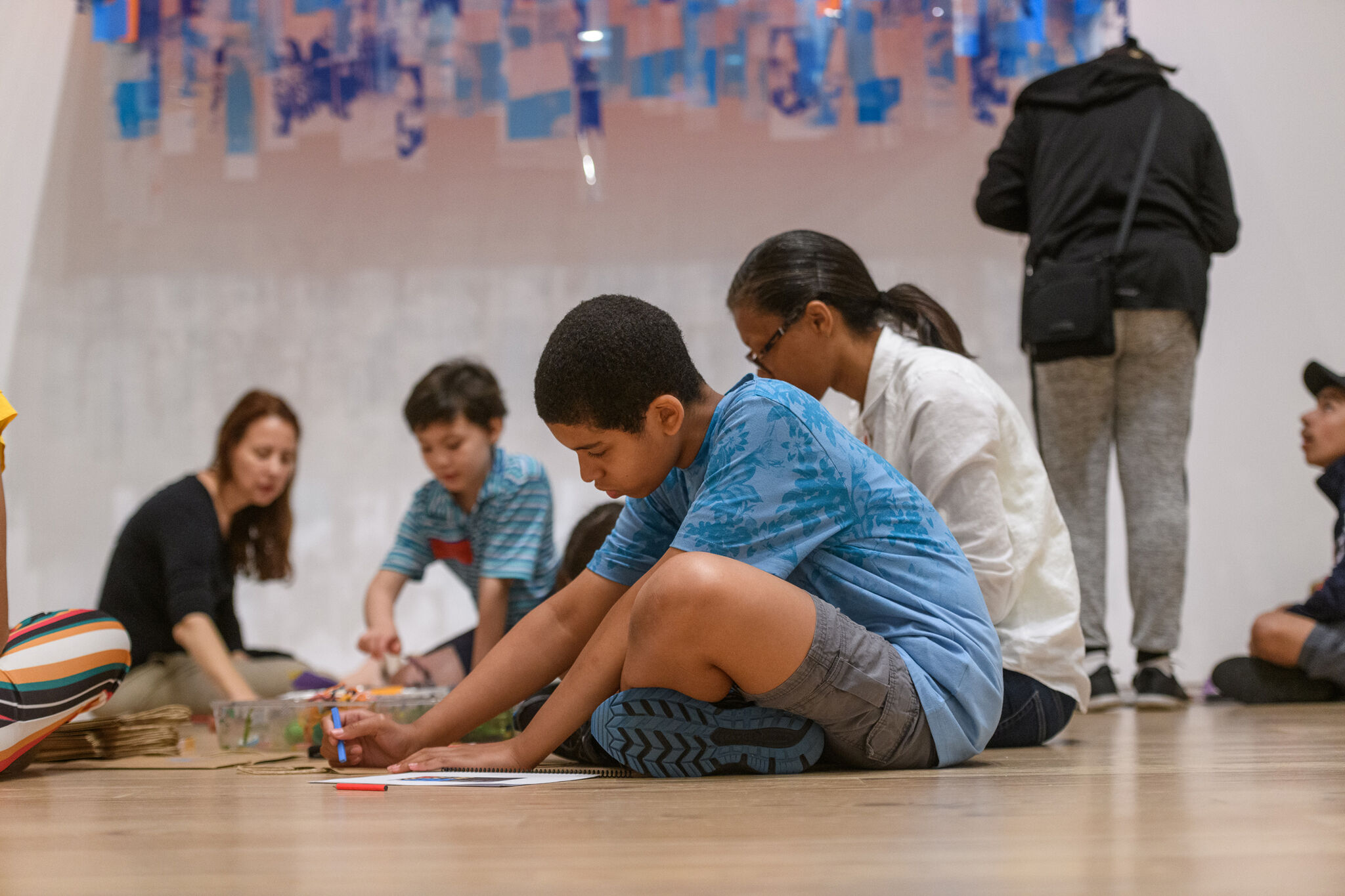 Adults and kids sit on the gallery floor, making art together.