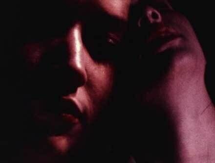 Dim close-up of two faces washed in deep purple tone. One leaned forward with shadows casting down the brow, nose, and lips, the other leaned back, neck lit from below.