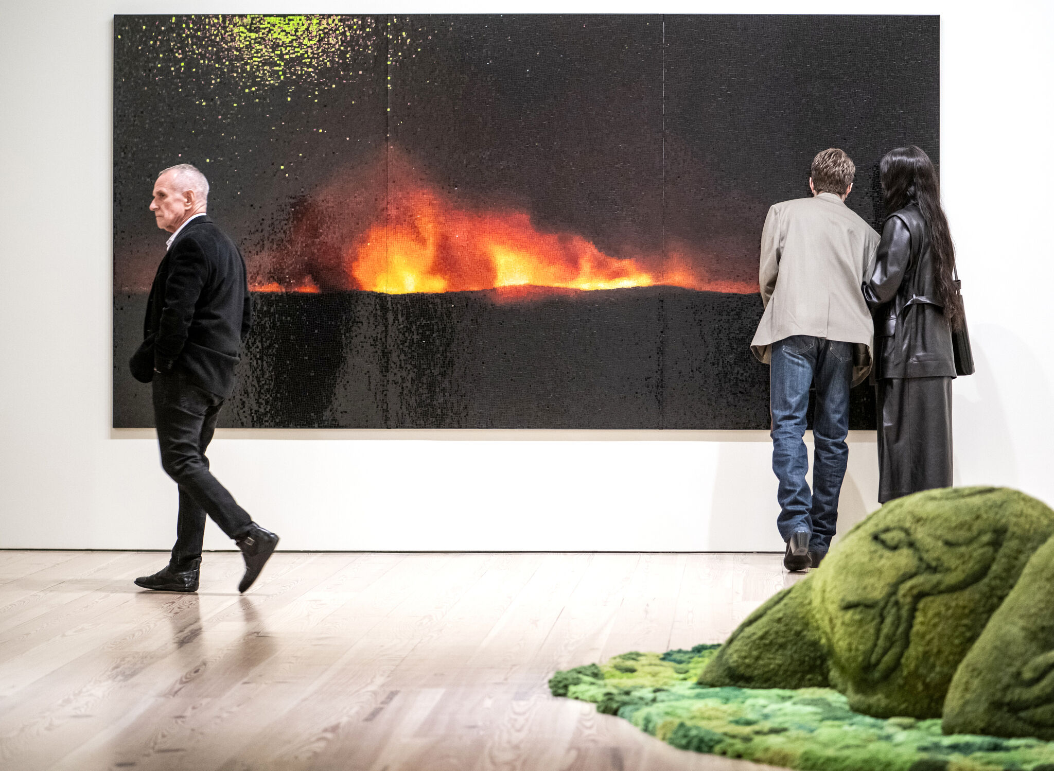 People gazing at a mosaic that looks like land on fire, behind a mossy sculpture on the ground with different shades of green.