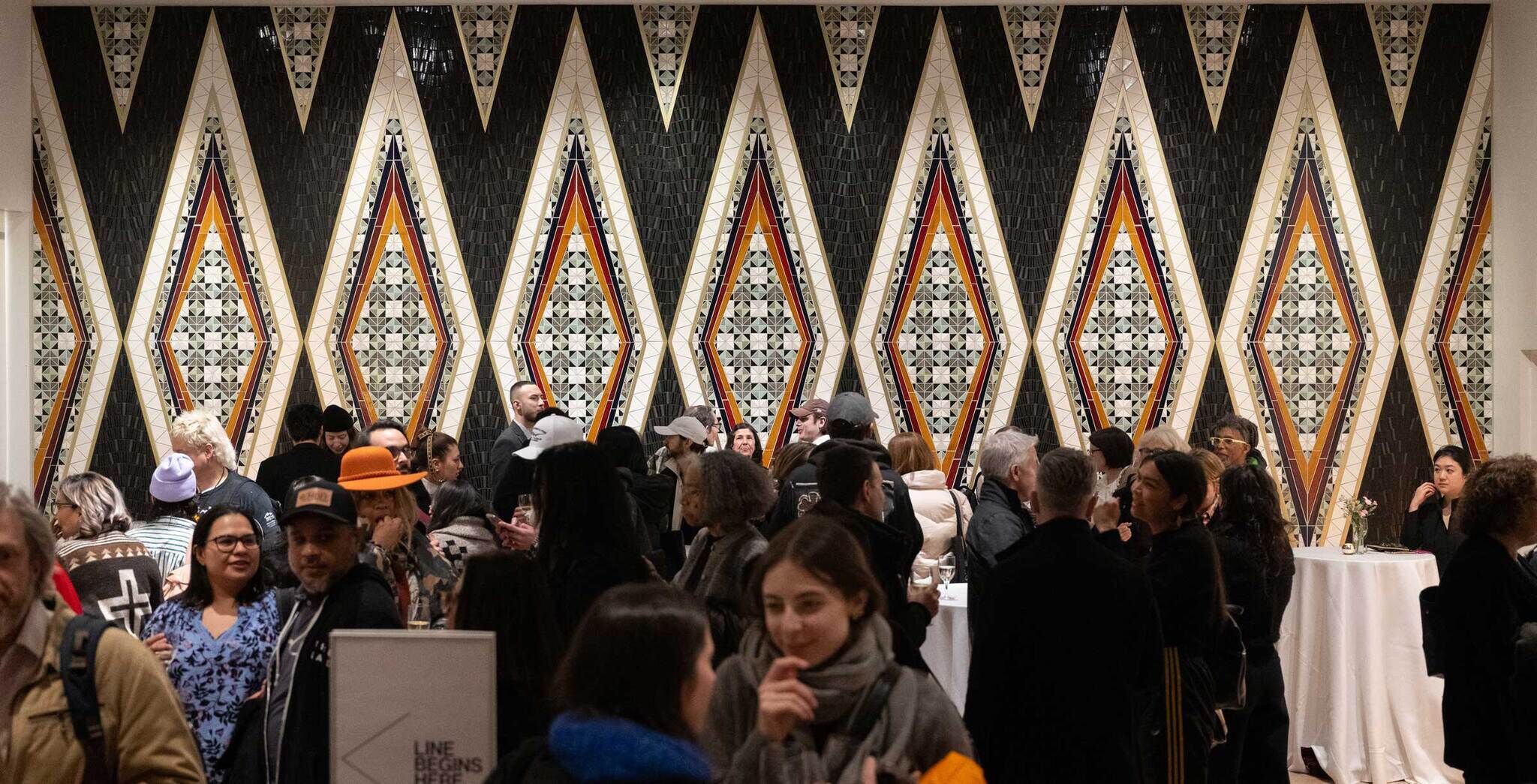 A crowded gallery reception with people mingling in front of a large geometric tile wall.