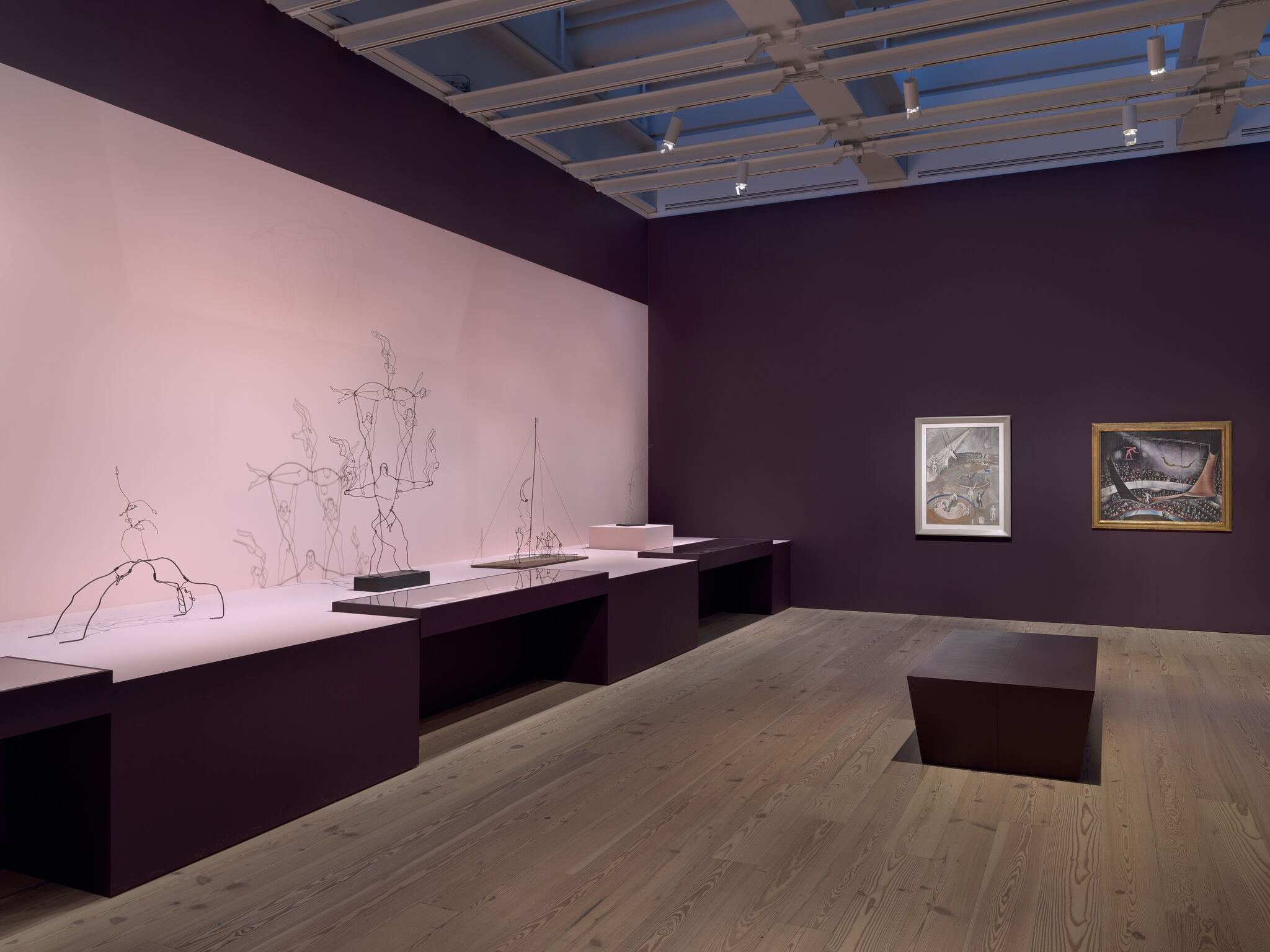 Wire sculptures on a long pink display table with two framed paintings on a dark purple wall.