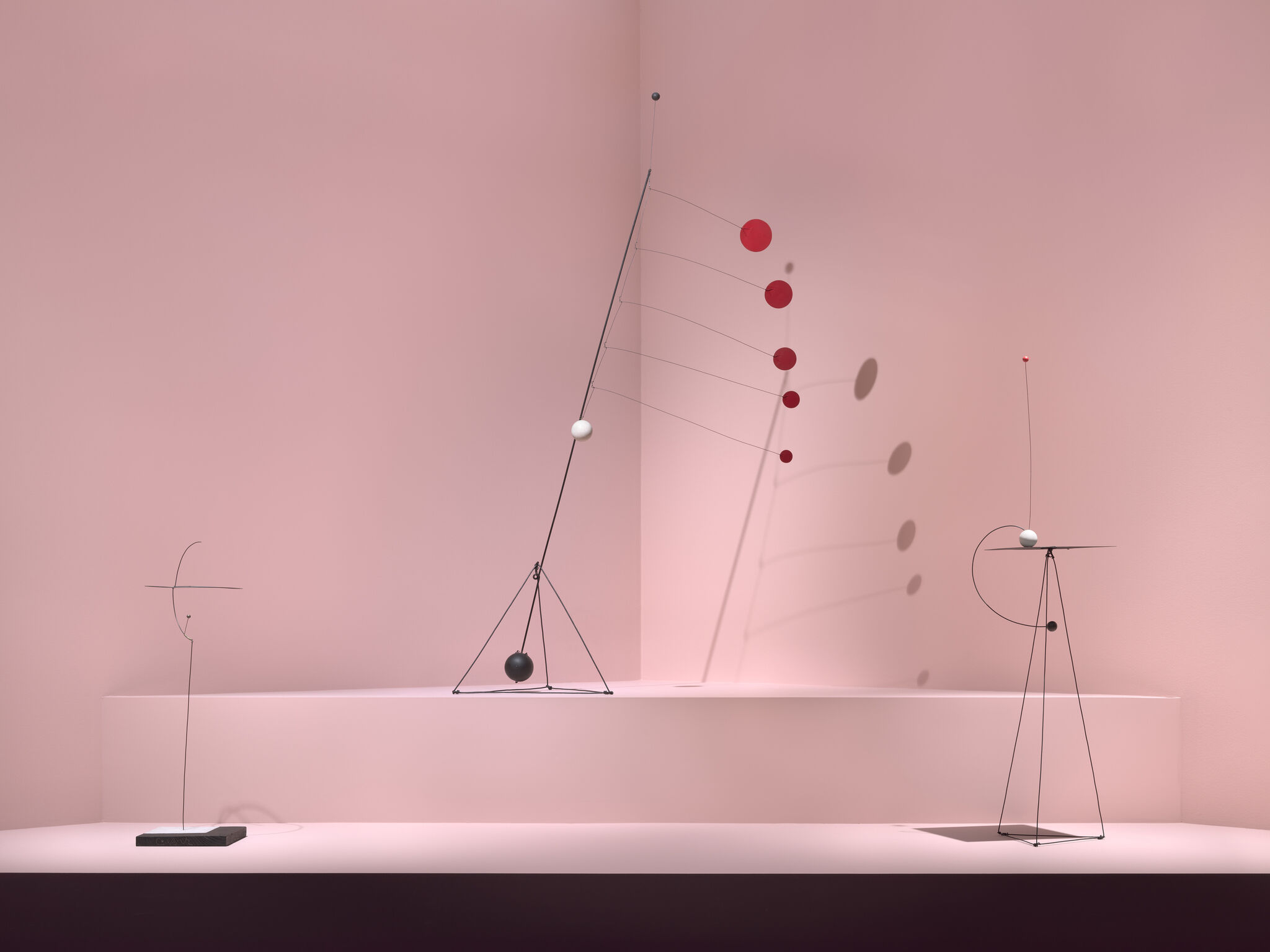 Three minimalist metal mobiles with hanging spheres and red discs casting shadows on a pink wall.