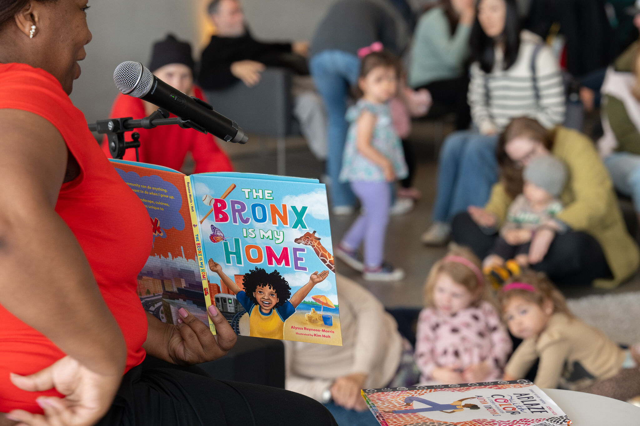 A Black woman in a red shirt holds a book titled "The Bronx is My Home" while reading aloud to a diverse group of youth and adults sitting cross-legged on the floor during the Second Sunday Storytime event.