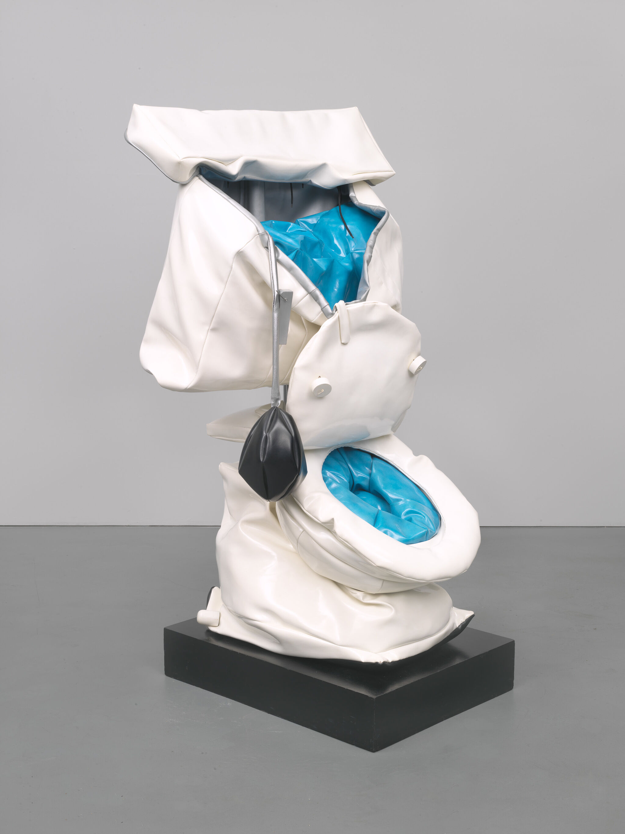 A white sculptural toilet with bright blue cushioned interiors shown in a flexible and folded state. A black pump-like element hangs from the side. The artwork is displayed on a black rectangular base against a plain gray background. 