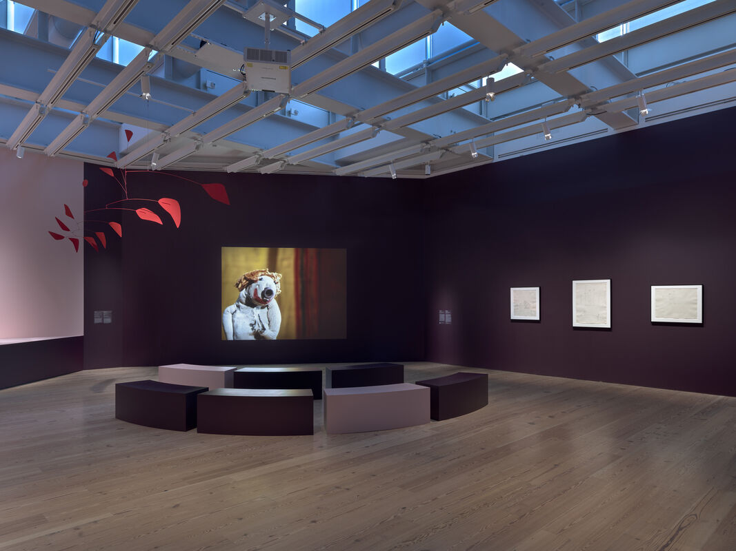 A museum gallery with curved seating and a projected puppet video on a dark purple wall.