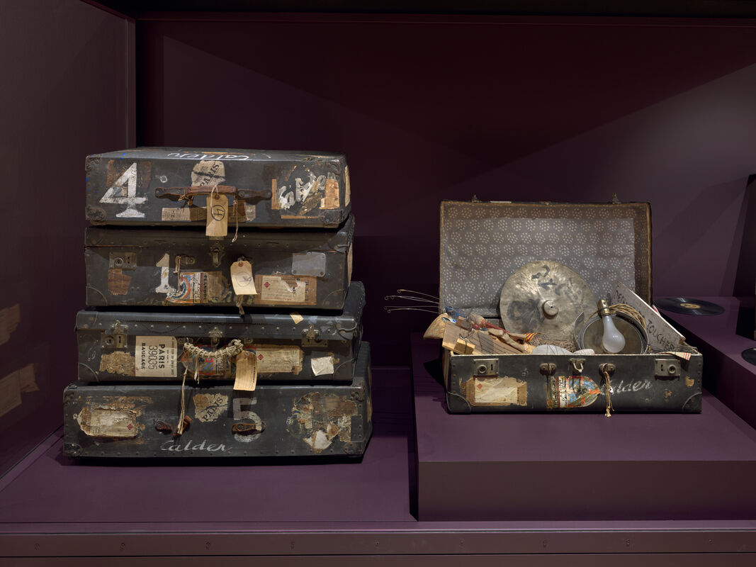 Stacked vintage suitcases with travel tags sit beside an open case holding cymbals and a light bulb.