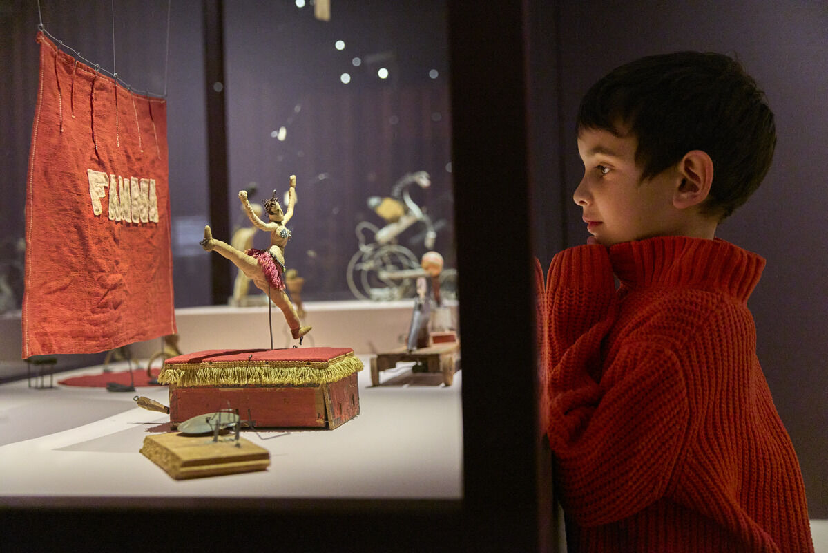 A boy in a red sweater watches a small toy acrobat mid-jump in a glass display.