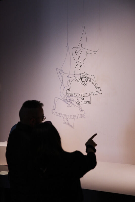 Two visitors point at a hanging wire sculpture labeled "Wire Sculpture by Calder."