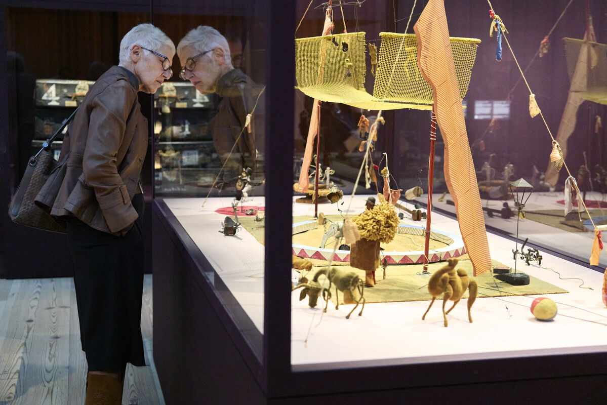 An older woman leans forward to closely examine a miniature circus display behind glass.