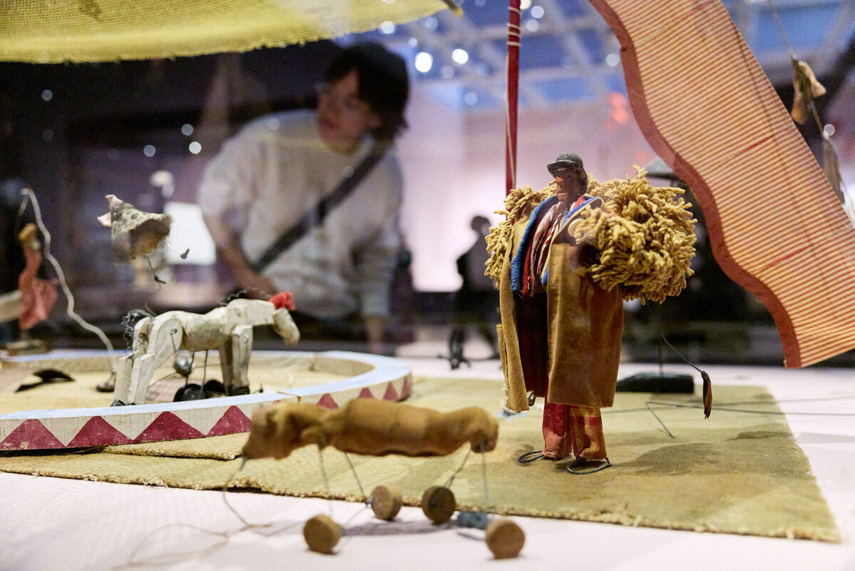 A small circus figurine wearing a shaggy coat stands beside toy animals on a miniature stage.