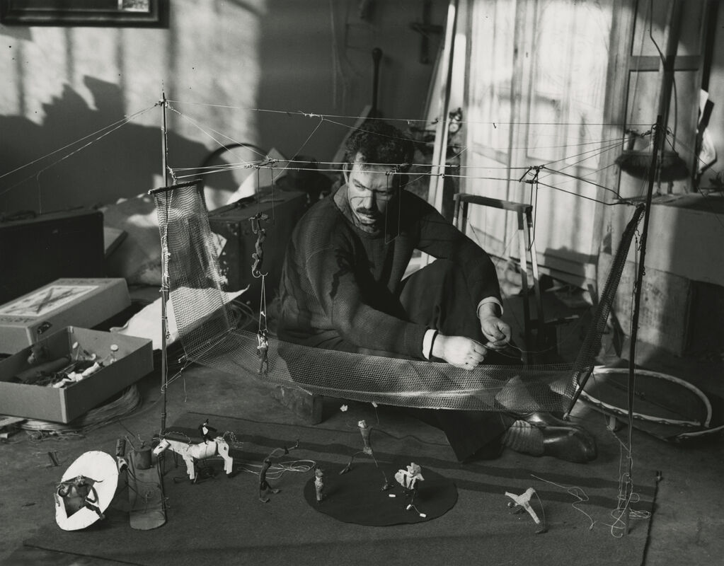 A man sits on the floor assembling a wire and mesh puppet stage with small figurines.