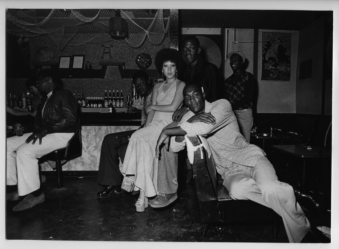 A black and white photograph of people in a dimly lit bar in Okinawa. The central grouping of figures includes four Black men gathered around an Okinawan woman who is wearing a long dress and an afro hairstyle.