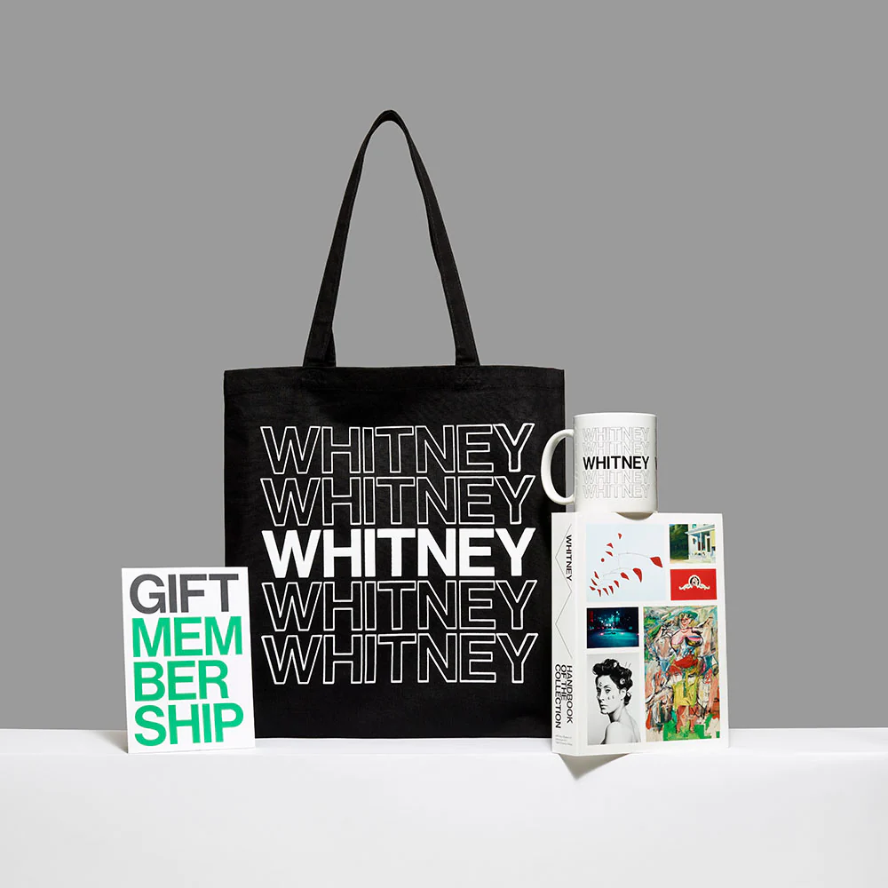 Black tote bag printed with repeated 'Whitney' text displayed alongside a mug, book, and gift membership card.