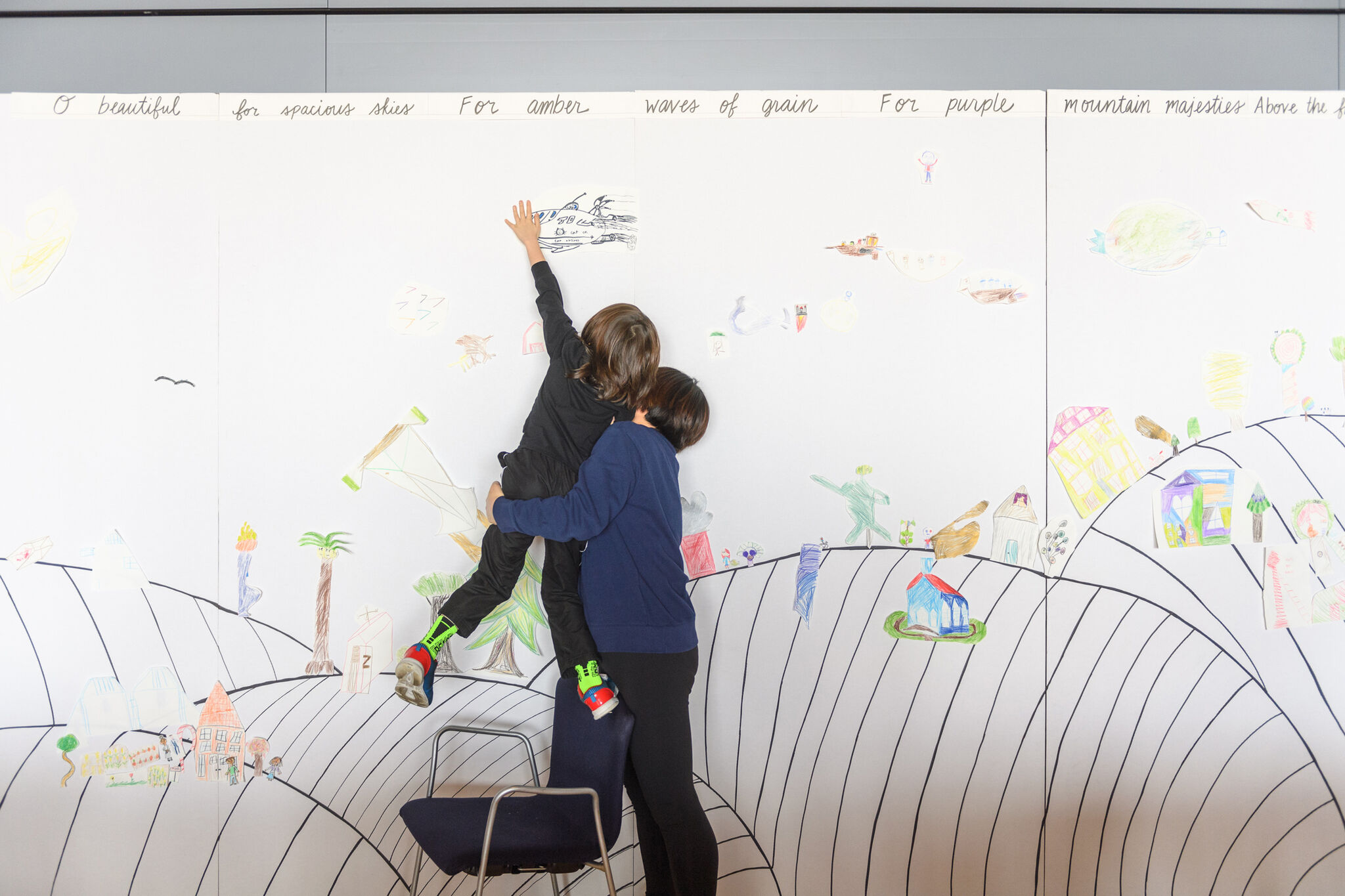 A child reaches up to draw on a large colorful drawing while an adult lifts and supports them.