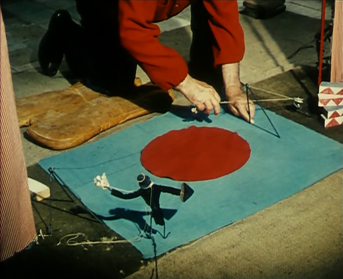 Person kneeling manipulates a small marionette puppet on a blue mat with a red circle.