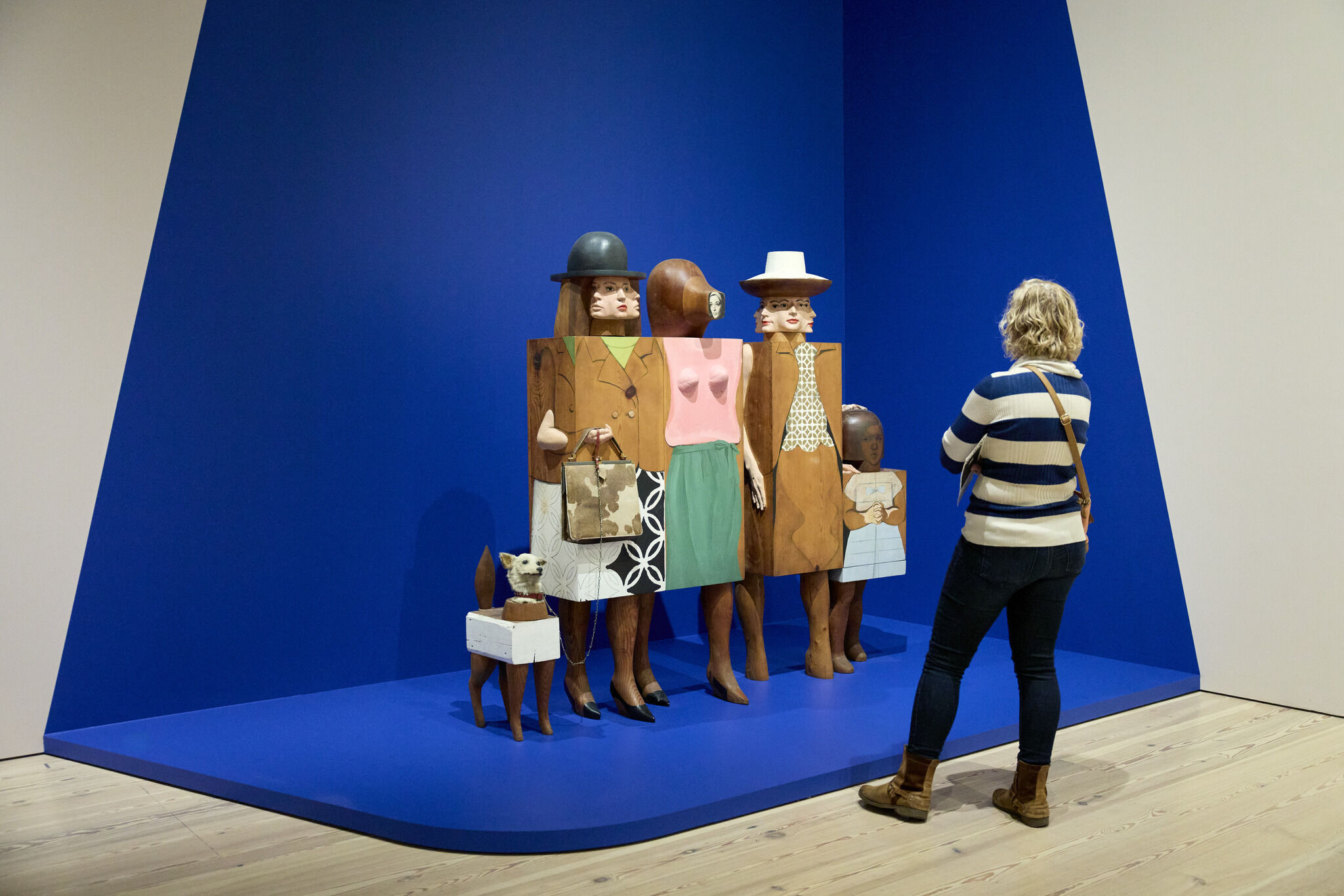 A woman with gray hair stands in front of a wooden sculpture of cubic women with a dog on a leash. 
