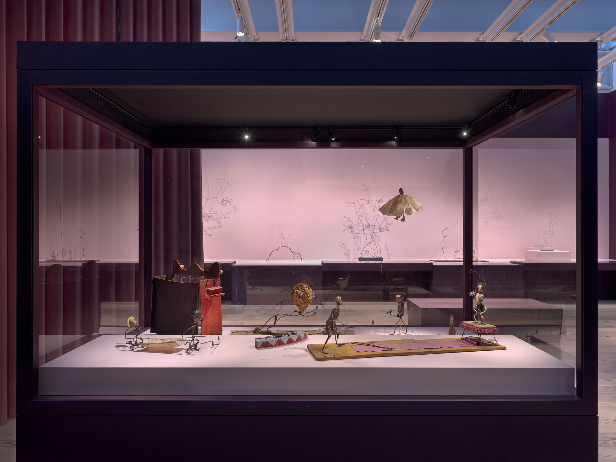 Glass display case shows small puppet-like sculptures arranged on a shallow stage.
