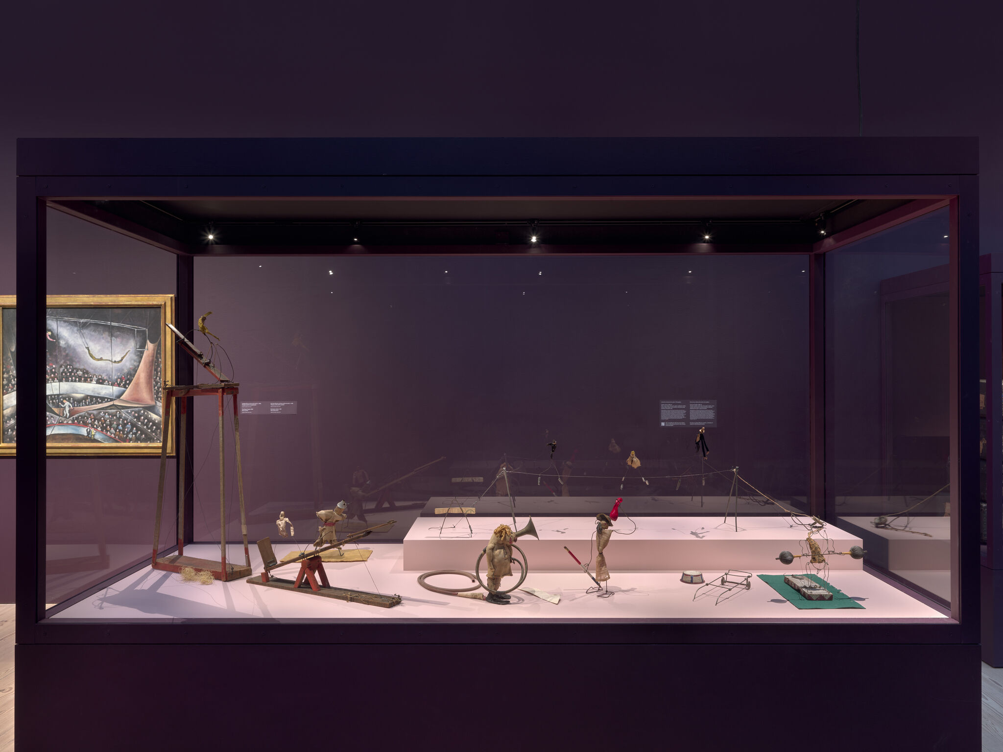 A glass museum case displays vintage toy puppets and mechanical figures arranged on a pink platform.