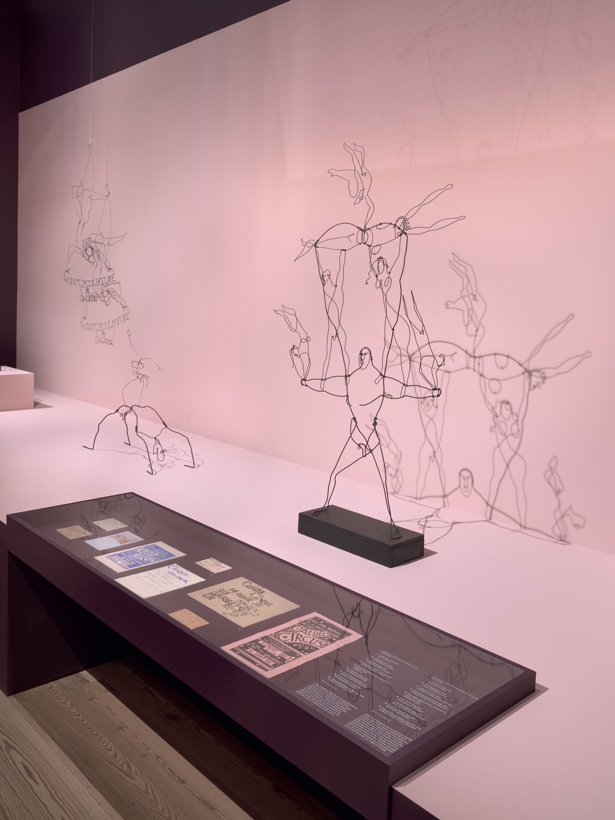 Delicate wire sculptures of acrobats cast shadows on a pink wall above a display case.