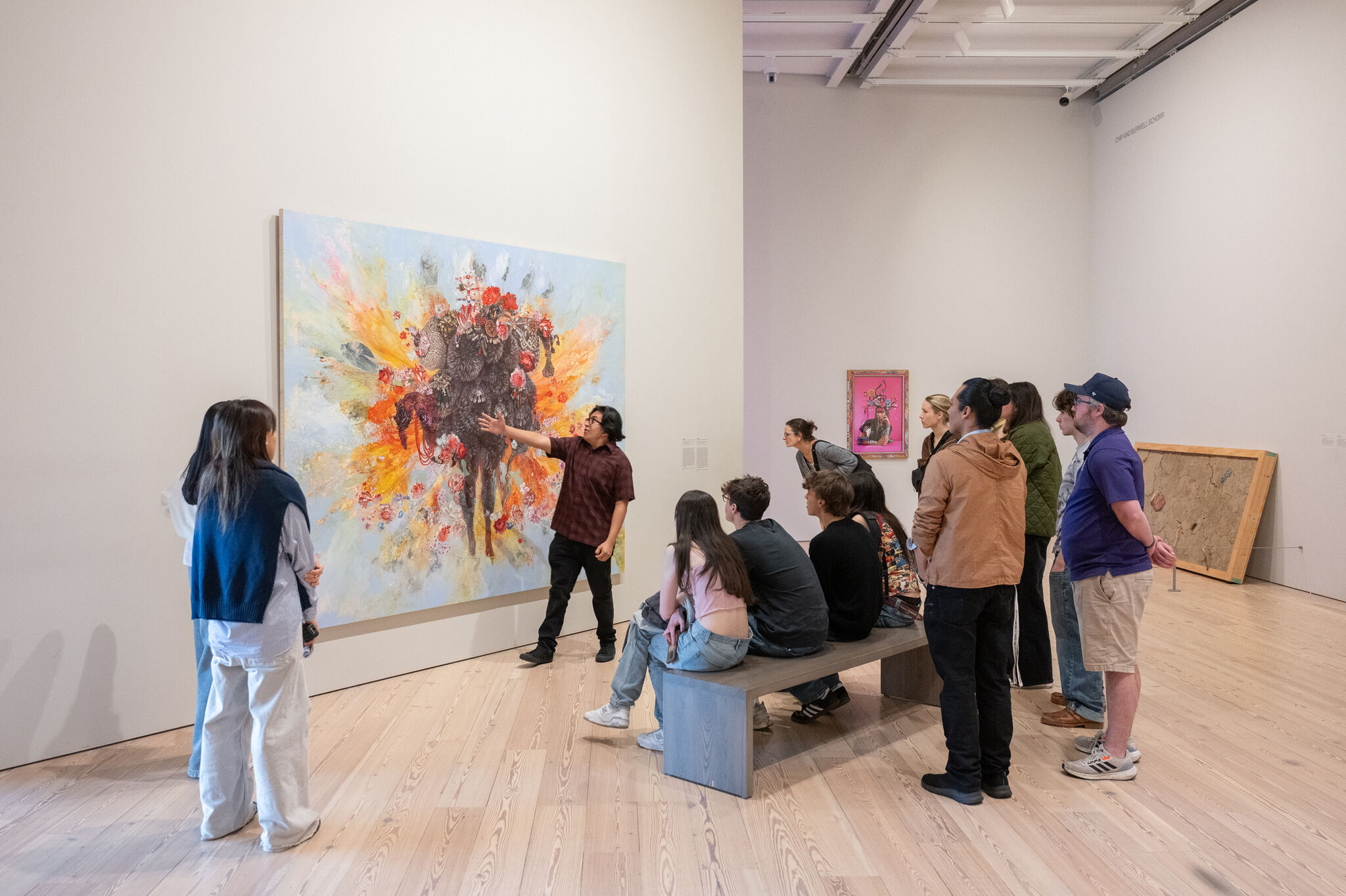 A museum guide points at a large colorful painting while a group of visitors listens.