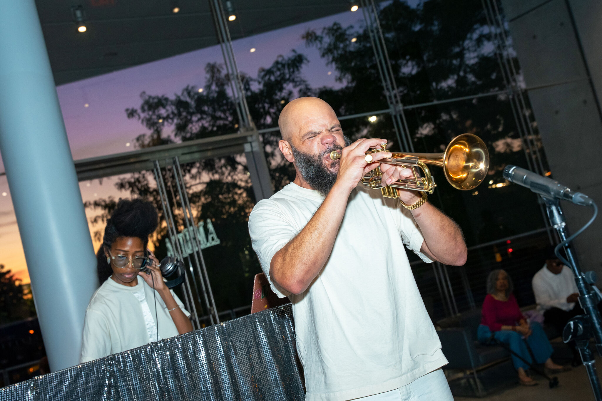 Musician passionately plays trumpet while DJ adjusts headphones behind him at evening event.