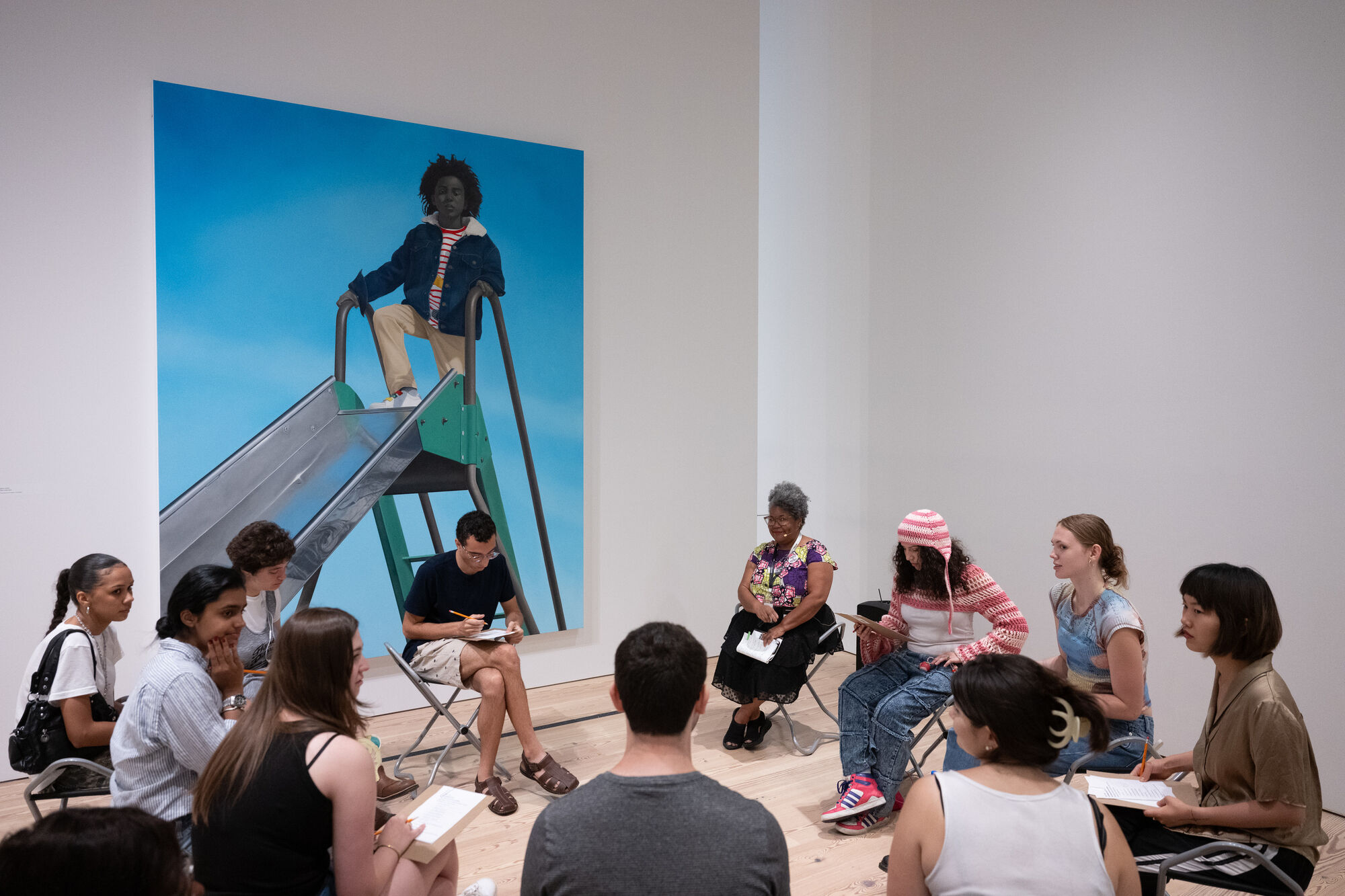 A diverse group sits in a museum discussion circle beneath a large portrait of a person on a slide.