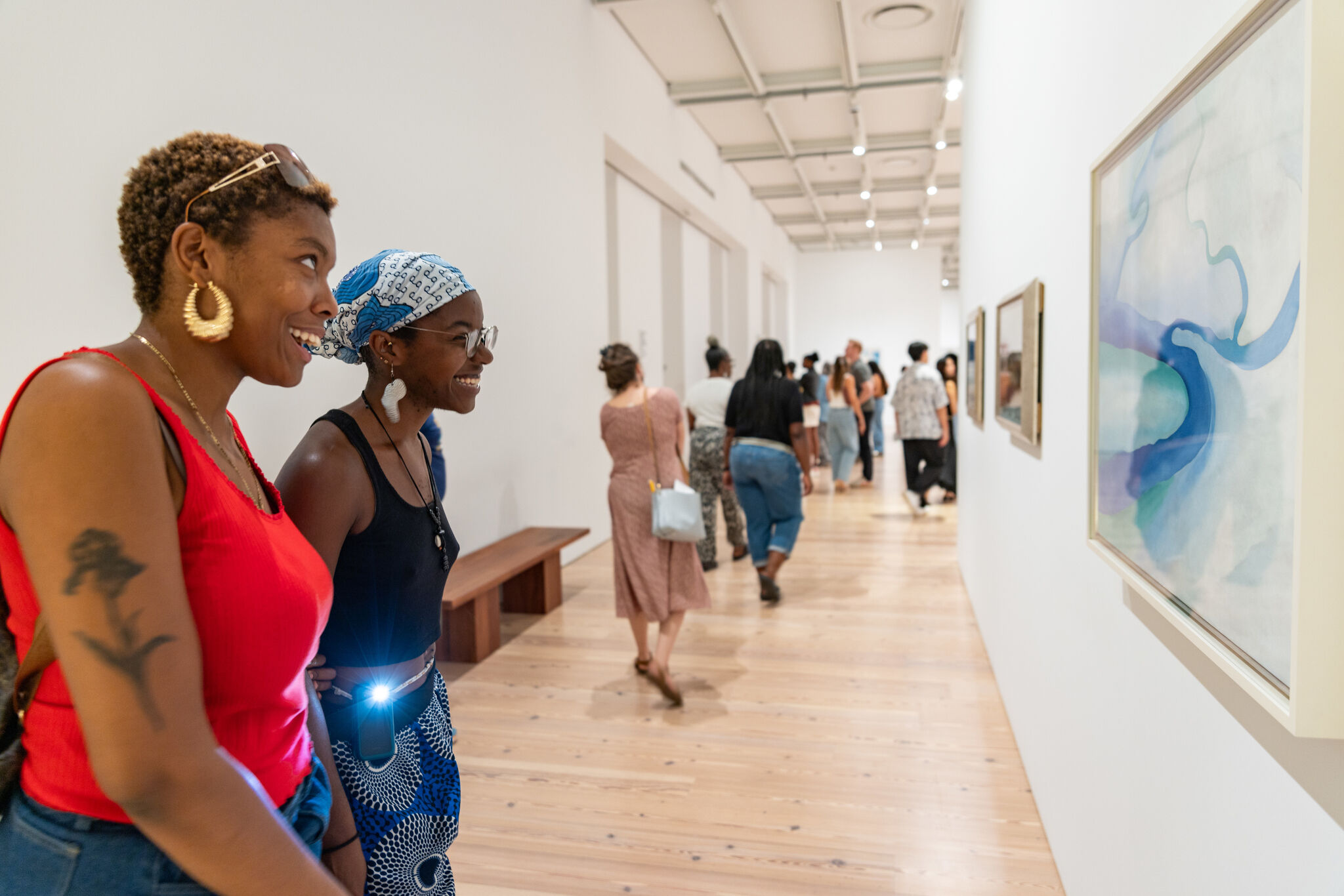 Two women smiling and admiring a painting while walking through a busy art gallery.
