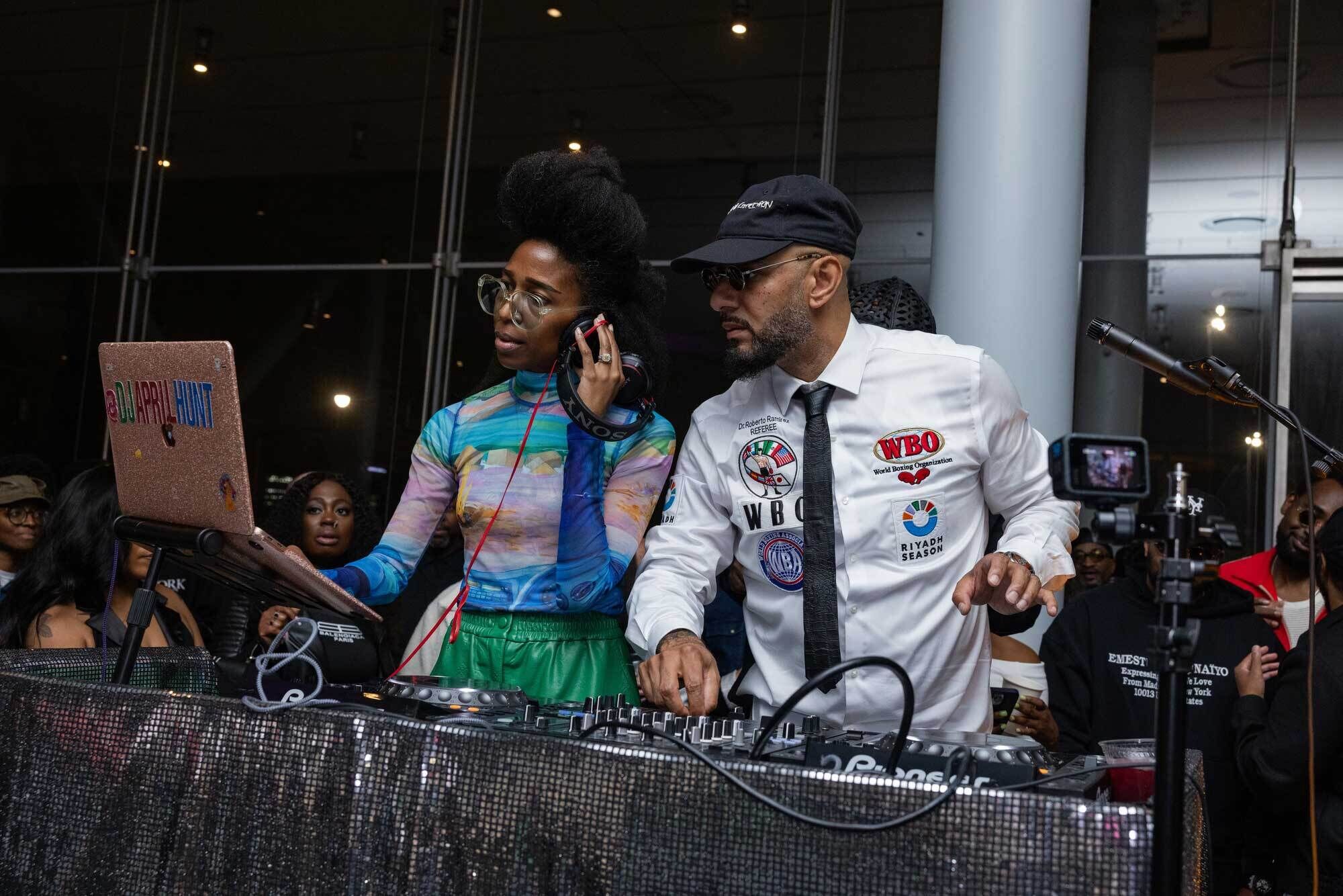 Two DJs collaborate at a crowded DJ booth as one listens on headphones and the other adjusts controls.