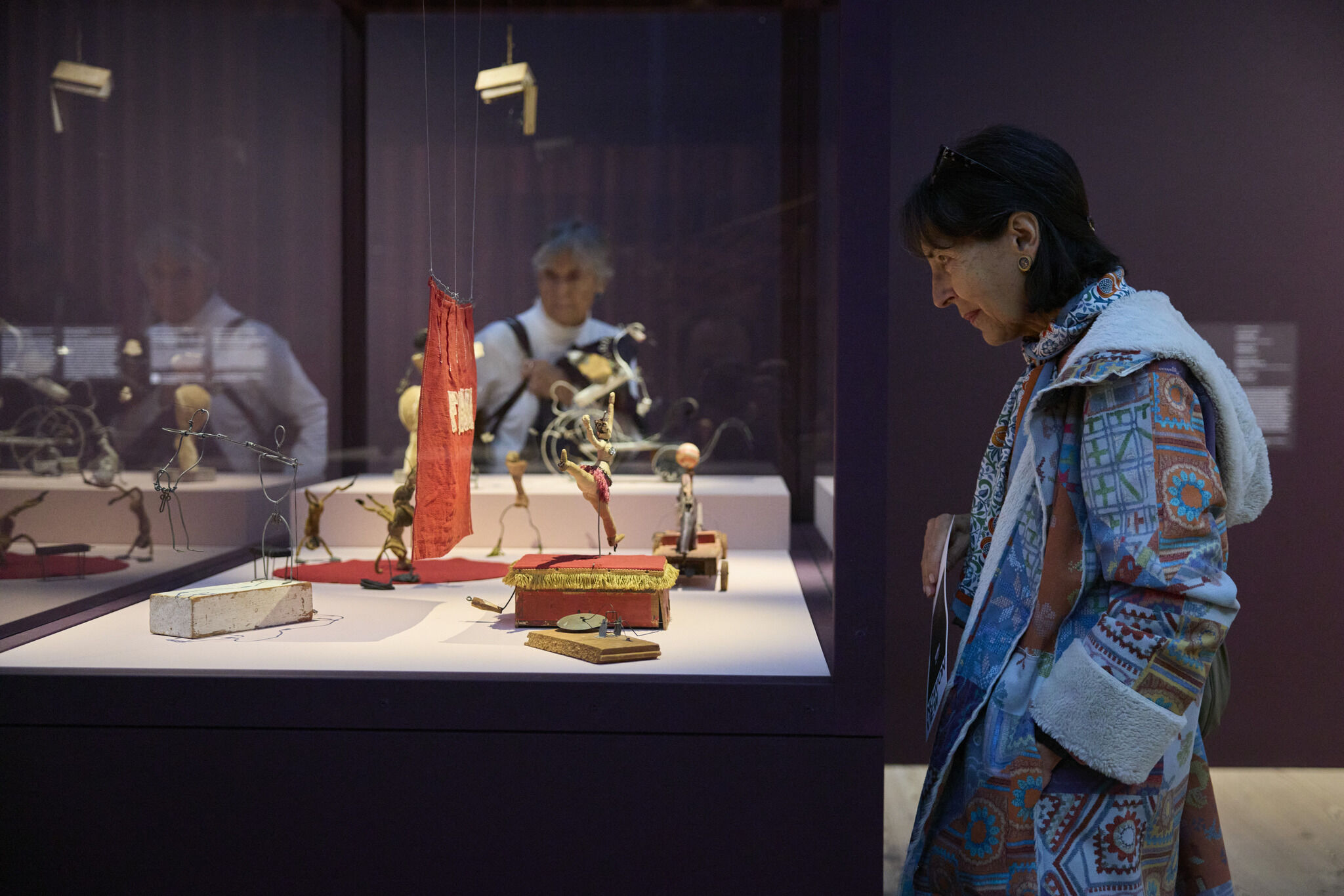 Middle aged woman smiles while gazing into a glass case of wire and fabric circus figures.