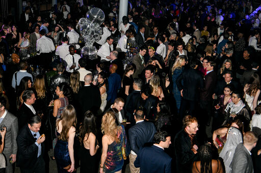 Large crowd of well-dressed people mingling and drinking under mirrored disco ball at a party.
