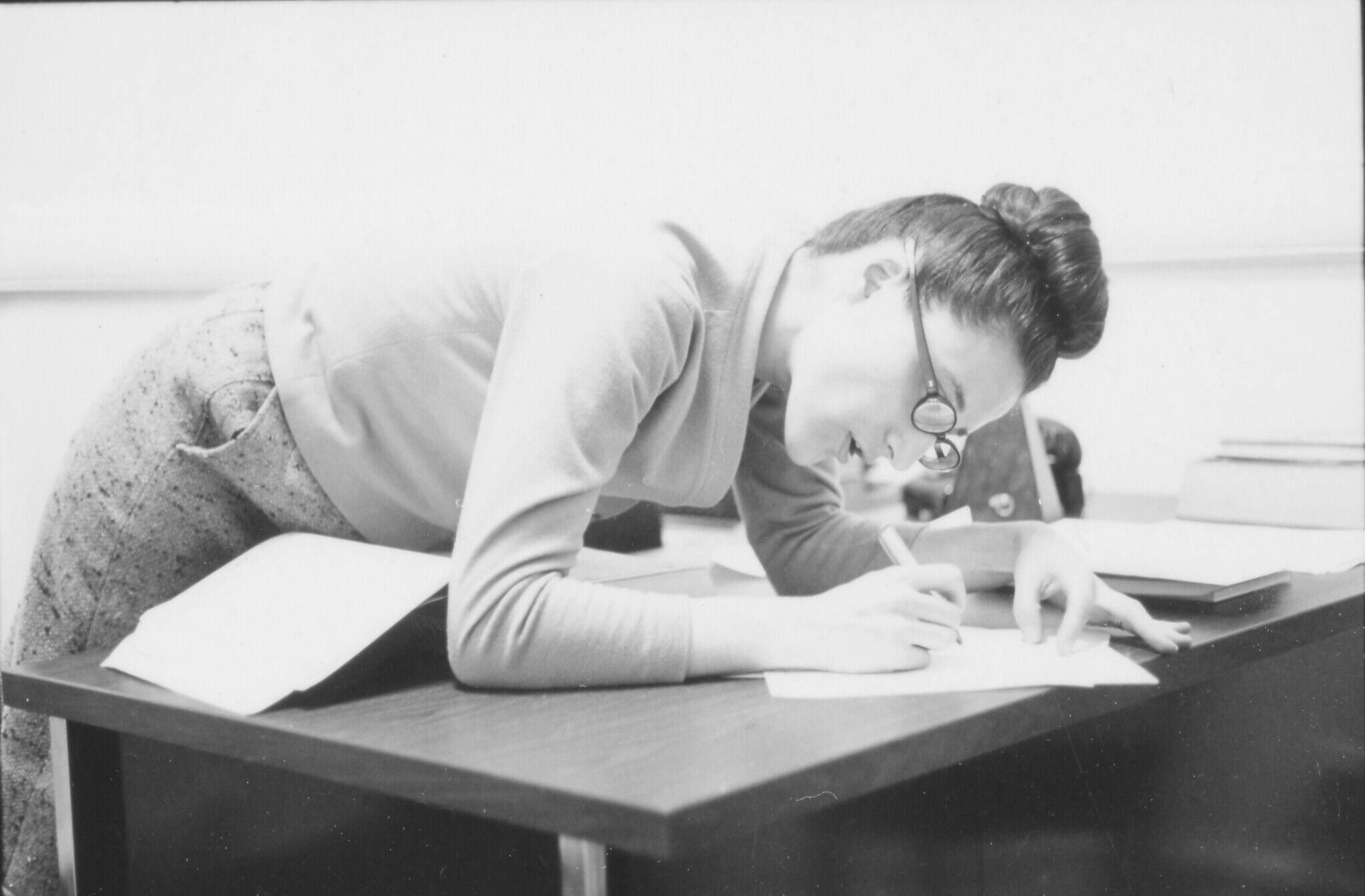 A black and white photograph of a white woman wearing glasses leaning over a wooden desk There are books and papers on the desk and the woman is writing on a piece of paper.  