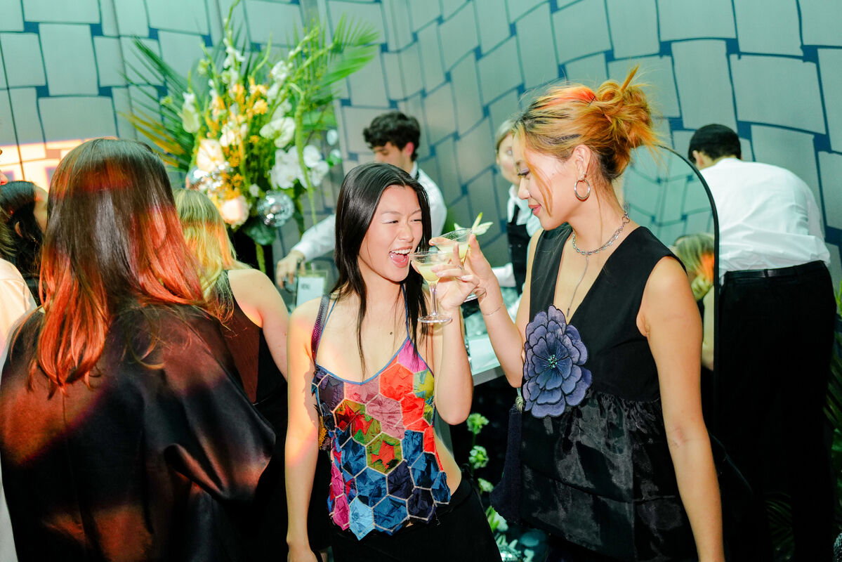 Two women clinking cocktail glasses and laughing at a lively party.