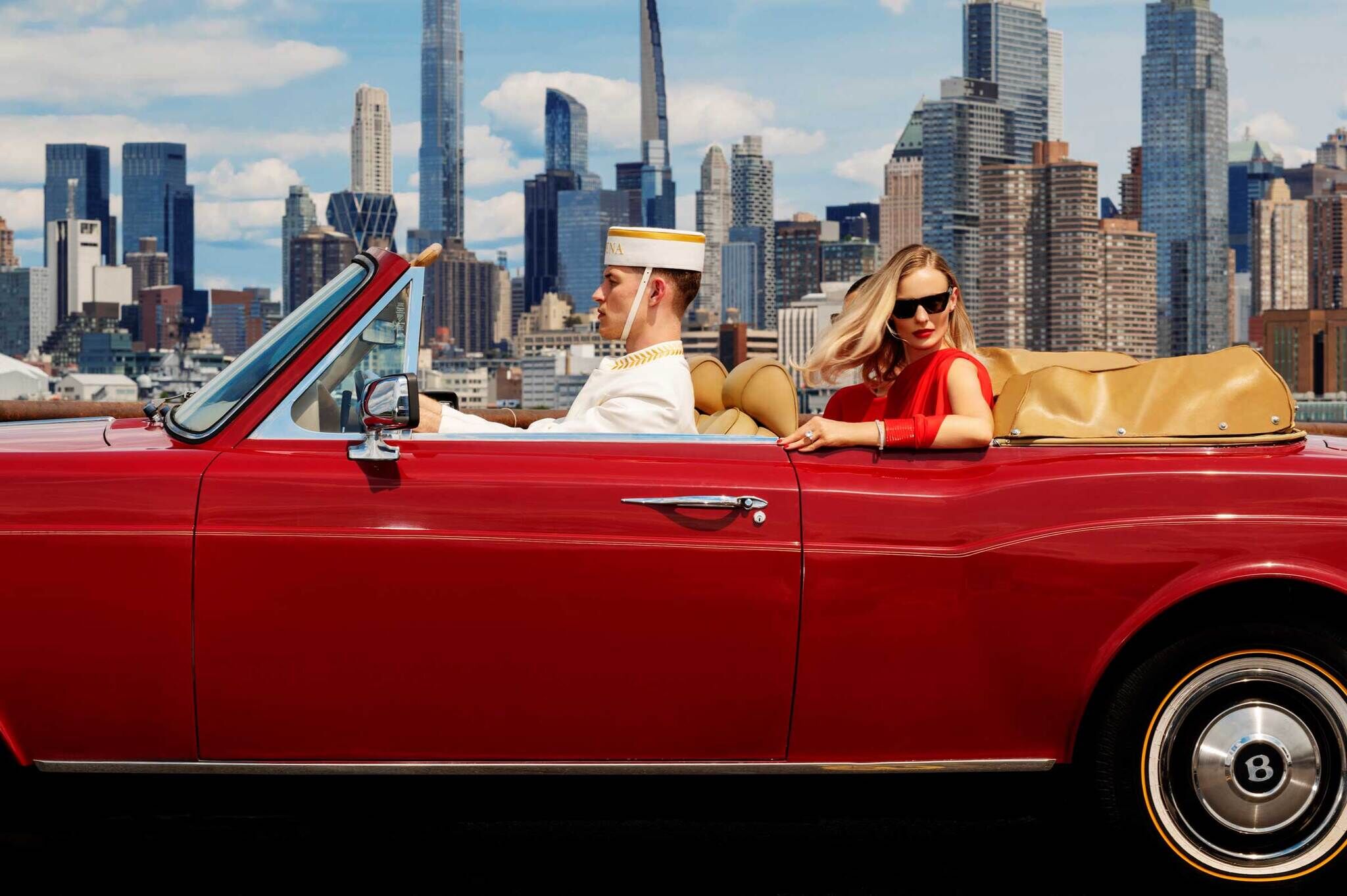 A woman in a red clothing and sunglasses looks out from the backseat of a red car. In the background is the NYC cityscape.