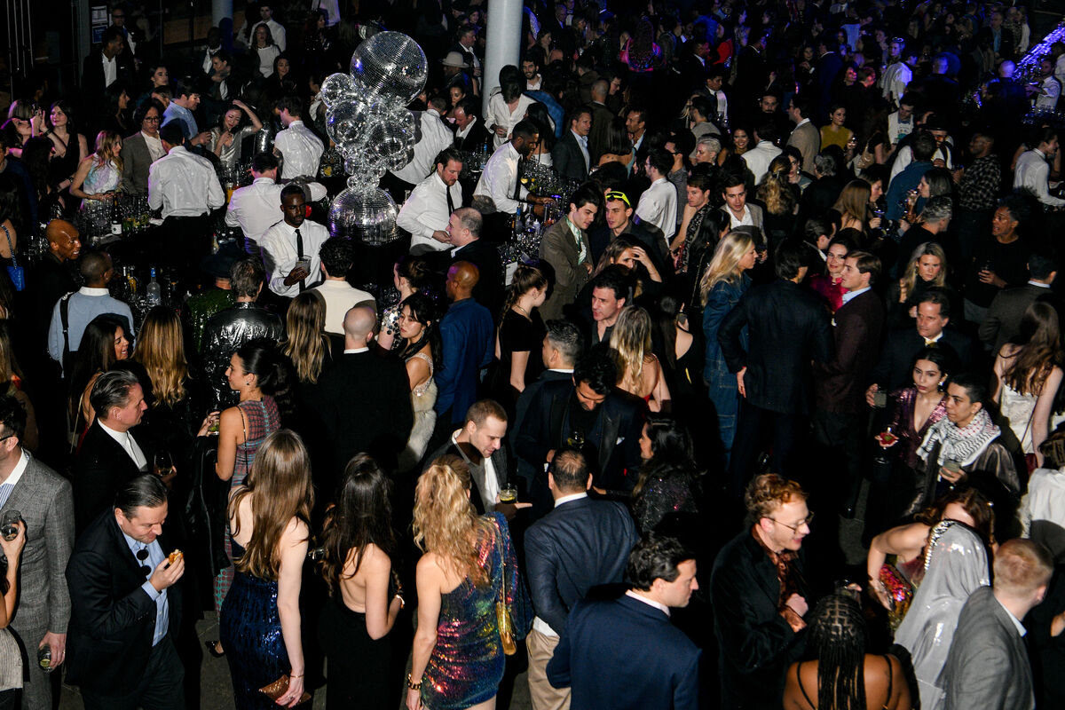 A crowded party with people mingling around a central bar and mirrored disco ball decorations.